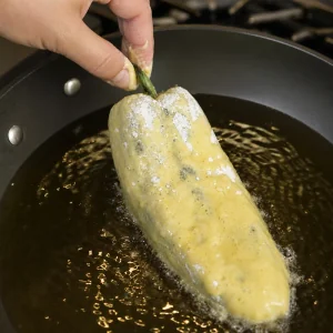 A battered stuffed poblano pepper lowered into hot oil in a skillet.