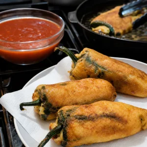 Golden fried chiles rellenos draining beside warm tomato sauce.