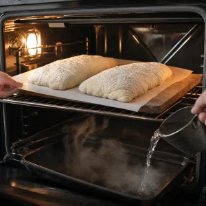 Ciabatta loaves slid onto a hot stone while water creates steam below