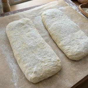 Two flour-dusted ciabatta loaves resting on parchment paper before baking