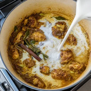 Coconut milk swirling into browned chicken curry at a gentle simmer.