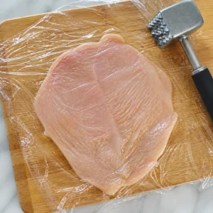 Thin chicken breast flattened under plastic wrap with a meat mallet nearby