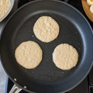 Tres porciones pequeñas de masa de panqueques de plátano separadas en una sartén antiadherente.
