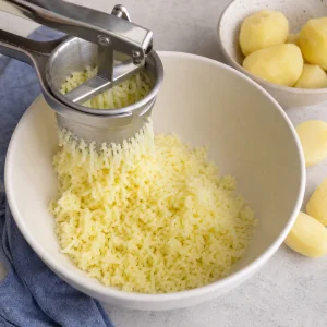 Cooked potatoes being pressed through a ricer into a mixing bowl