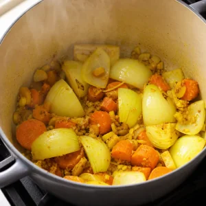 Quartered onion softening with ginger, turmeric, garlic, and carrots in a soup pot