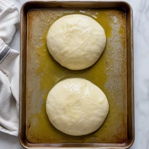 Two focaccia dough pieces resting on an oiled baking sheet