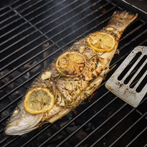 Grilled whole fish flipped on grill grates with lemon slices and crushed garlic on top
