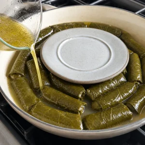 Бульон и оливковое масло наливают поверх долмы, маленькая тарелка удерживает рулеты под жидкостью