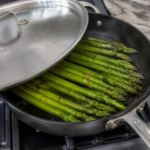 Espárragos cocinándose en aceite de oliva bajo una tapa entreabierta