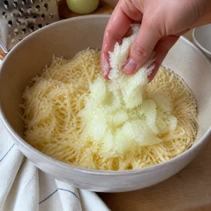 Grated onion added to a bowl of drained potatoes