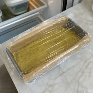 Loaf pan of pistachio tahini halva covered with plastic wrap before chilling