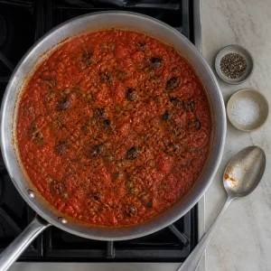 Simmered puttanesca sauce with a tasting spoon, salt, and black pepper nearby.