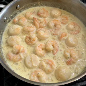 Garnelen und Jakobsmuscheln köcheln sanft in heller Cremesauce in einem Topf.