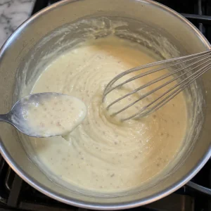 Glatte Milch-Weißwein-Sahnesauce dickt in einem Topf mit Schneebesen und Löffel ein.